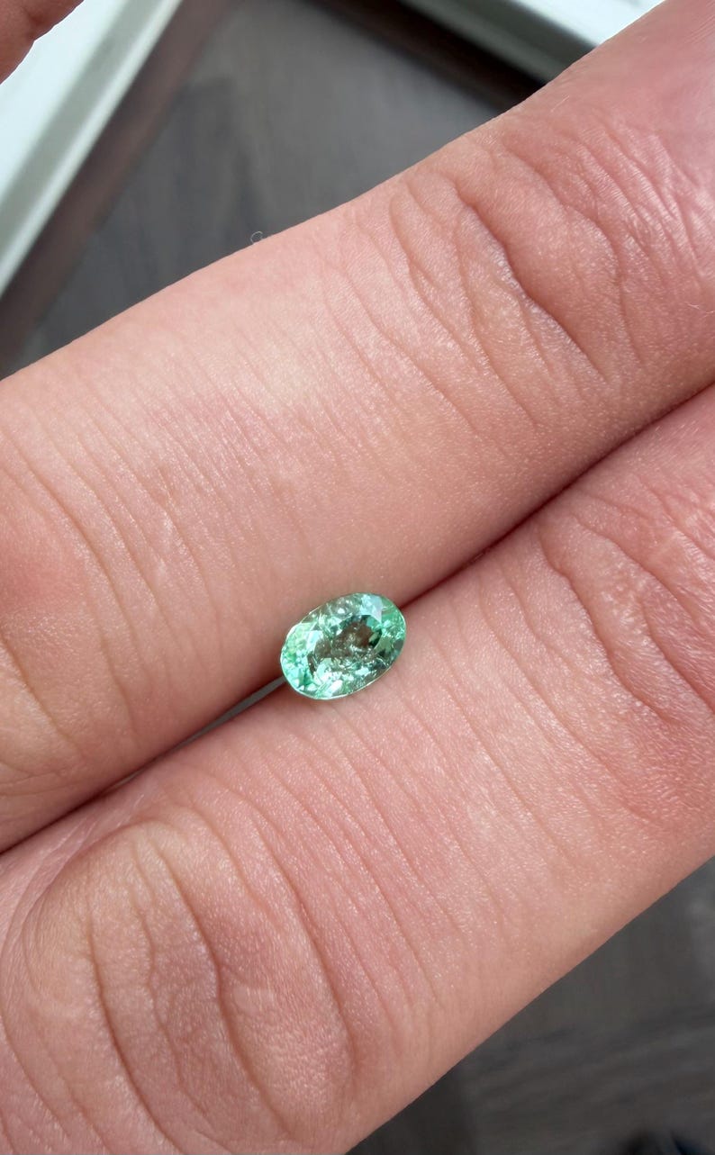May include: A small, oval-shaped, light green gemstone with internal inclusions. The gem is held between fingers, showcasing its color and clarity. The gemstone is likely an emerald, a popular choice for jewelry.