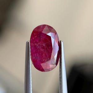May include: An oval-shaped, faceted ruby gemstone with a deep red color and visible internal inclusions, held securely by a pair of silver-toned metal tweezers. The gemstone displays a polished surface with multiple geometric facets reflecting light.