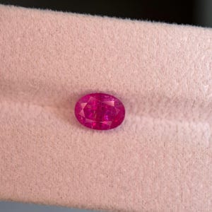 May include: A vibrant, oval-cut gemstone with a deep magenta-pink hue and a faceted surface that reflects light. The stone displays a clear, transparent quality with multiple geometric facets across its top, showcasing a brilliant, polished finish.
