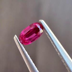 May include: A vibrant, oval-cut ruby gemstone, held by silver tweezers. The ruby displays a deep, saturated red hue with internal facets reflecting light. The gemstone's shape is elongated, with rounded ends, and is set against a neutral background.