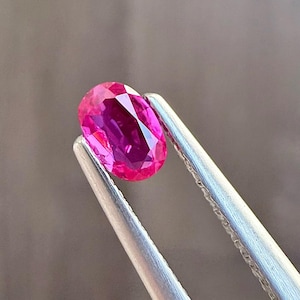 May include: A vibrant, oval-cut ruby gemstone held by silver tweezers. The ruby displays a deep pinkish-red hue with internal facets reflecting light. The tweezers are positioned against a blurred, neutral background, highlighting the gemstone's brilliance.