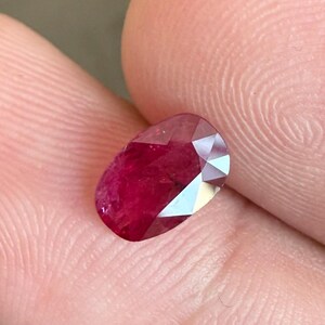 Puede incluir: Una gema de rubí facetada, de forma ovalada. El rubí es de un color rojo intenso y saturado con múltiples facetas que reflejan la luz. La gema se sostiene entre dos dedos, mostrando su tamaño y brillo. El fondo es de un tono neutro.
