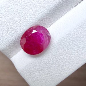 Può includere: Una gemma di rubino di forma ovale con una superficie sfaccettata che riflette la luce. La pietra rosso-rosata profonda presenta inclusioni interne naturali e una finitura lucida, mostrando una brillante saturazione del colore in tutta la gemma.