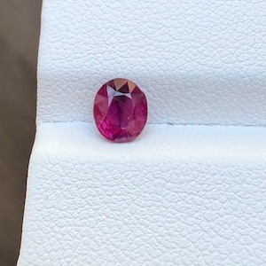 May include: Oval-cut, faceted ruby gemstone. The gem is a deep, vibrant red with internal light reflections. It is set against a textured white surface, showcasing the stone's brilliance. The ruby is a precious stone.