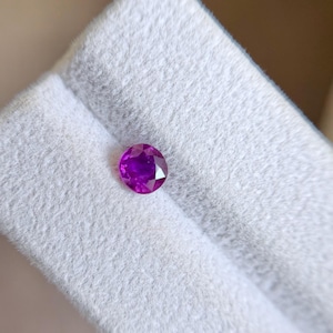 May include: A round, faceted gemstone with a vibrant magenta-pink hue. The stone features a brilliant cut with multiple triangular facets that reflect light, creating a sparkling appearance. It is positioned on a light grey, textured surface.