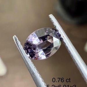 May include: An oval-cut, light purple gemstone is held by tweezers. The gem is faceted, reflecting light. Text indicates the gem weighs 0.76 ct and measures 5.2x6.91x3 mm. The background is a blurred, neutral tone.