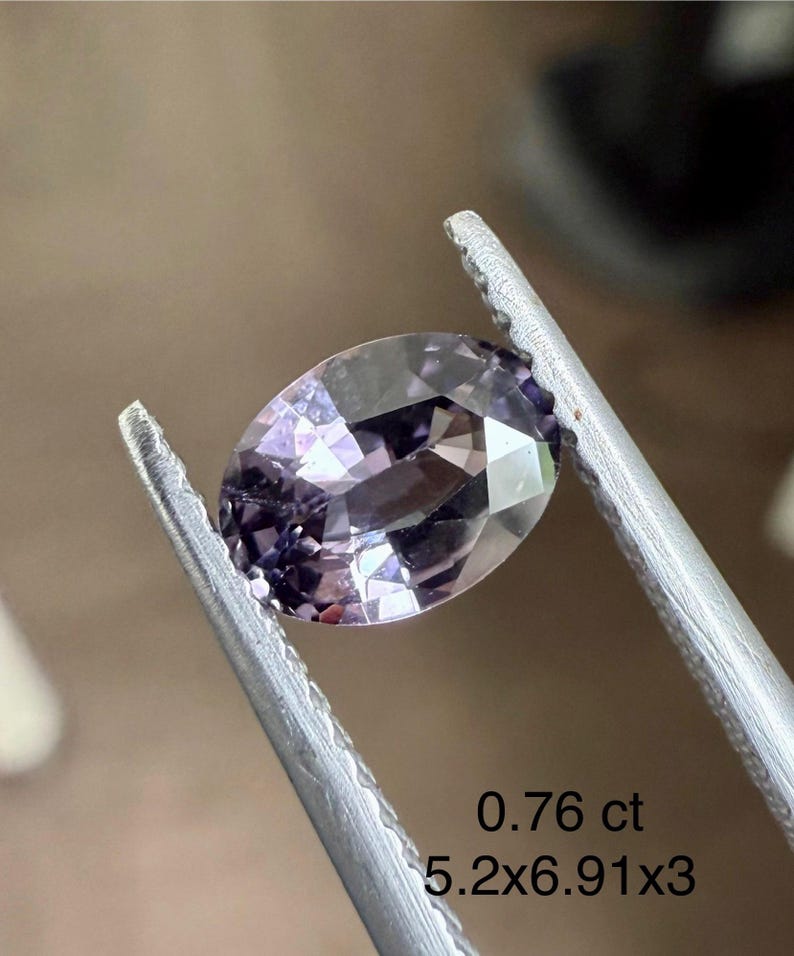 May include: An oval-cut, light purple gemstone is held by tweezers. The gem is faceted, reflecting light. Text indicates the gem weighs 0.76 ct and measures 5.2x6.91x3 mm. The background is a blurred, neutral tone.