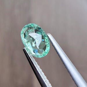 May include: A faceted, oval-shaped gemstone in shades of green and blue is held by a pair of silver tweezers. The gem has a brilliant cut, reflecting light and creating a sparkling effect. The background is a soft, blurred neutral color.