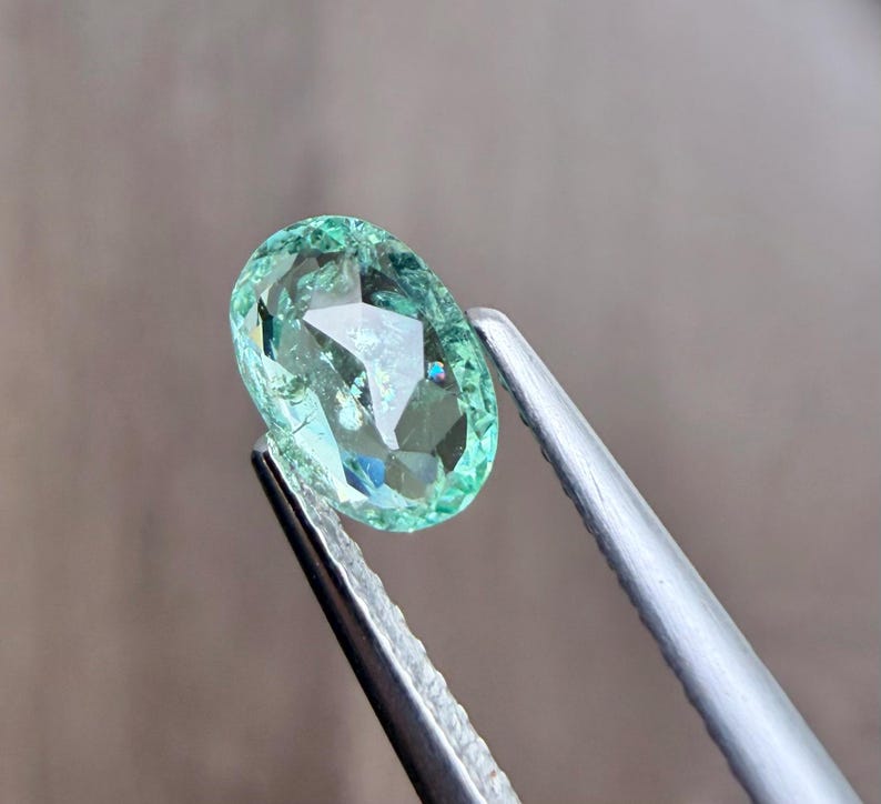 May include: A faceted, oval-shaped gemstone in shades of green and blue is held by a pair of silver tweezers. The gem has a brilliant cut, reflecting light and creating a sparkling effect. The background is a soft, blurred neutral color.