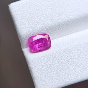 May include: A vibrant, cushion-cut pink gemstone with a faceted surface that reflects light. The stone displays a deep, saturated magenta hue and is positioned on a textured white surface, showcasing its clear, translucent quality.