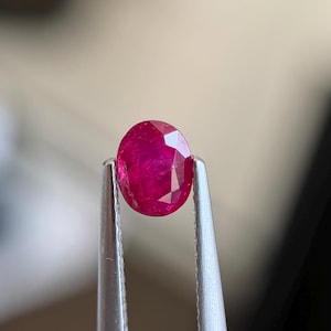 Könnte beinhalten: Ein leuchtend roter, oval geschliffener Rubin mit facettierter Oberfläche, der sicher zwischen den beiden silberfarbenen Metallspitzen einer Pinzette gehalten wird. Der Edelstein zeigt innere Klarheit und Lichtreflexionen.