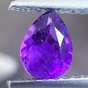 May include: A faceted, pear-shaped amethyst gemstone held by a metal tool. The gem displays a deep purple hue with internal inclusions and facets that catch the light. The stone's cut highlights its brilliance and clarity.