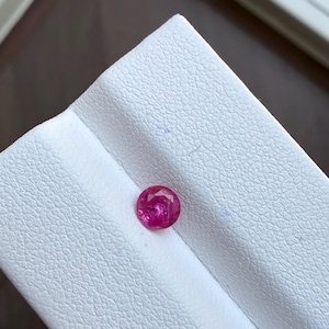 May include: A round, faceted, bright red gemstone. The ruby is set against a white textured surface. The gemstone is a vibrant, eye-catching color, and the facets create a play of light and shadow.