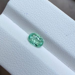 May include: An oval-shaped, light green gemstone. The gem is set against a white, textured surface. The gemstone has a slightly translucent appearance, with visible internal patterns and a polished surface. The overall color is a soft, inviting green.