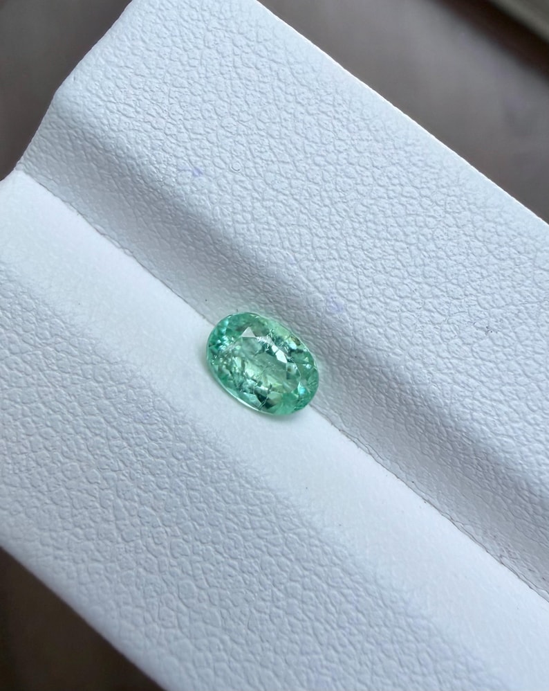 May include: An oval-shaped, light green gemstone. The gem is set against a white, textured surface. The gemstone has a slightly translucent appearance, with visible internal patterns and a polished surface. The overall color is a soft, inviting green.