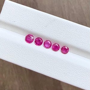 May include: Six round, faceted, vibrant red rubies are displayed in a white tray. The gemstones are arranged in a row, showcasing their polished surfaces and rich color. The rubies vary slightly in size and are set against a neutral background.