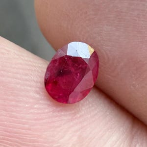May include: Oval-cut ruby gemstone with a deep red hue. The faceted surface reflects light, showcasing the gem's brilliance. The ruby is held against a skin-toned background, highlighting its color and shape. A precious stone.