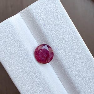 Könnte beinhalten: Ein facettierter, rund geschliffener Rubin-Edelstein. Der Rubin hat eine tiefe, leuchtende rote Farbe mit sichtbaren inneren Einschlüssen. Der Edelstein liegt auf einer weißen, strukturierten Oberfläche, die seine Farbe und Brillanz hervorhebt. Der Edelstein ist wahrscheinlich für Schmuck.