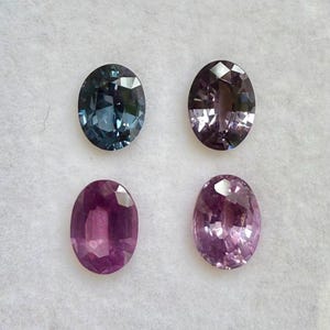 May include: Four oval-cut gemstones in shades of blue, purple, and pink are displayed against a white background. The gems vary in color intensity, with facets catching the light. These gemstones are suitable for jewelry making.