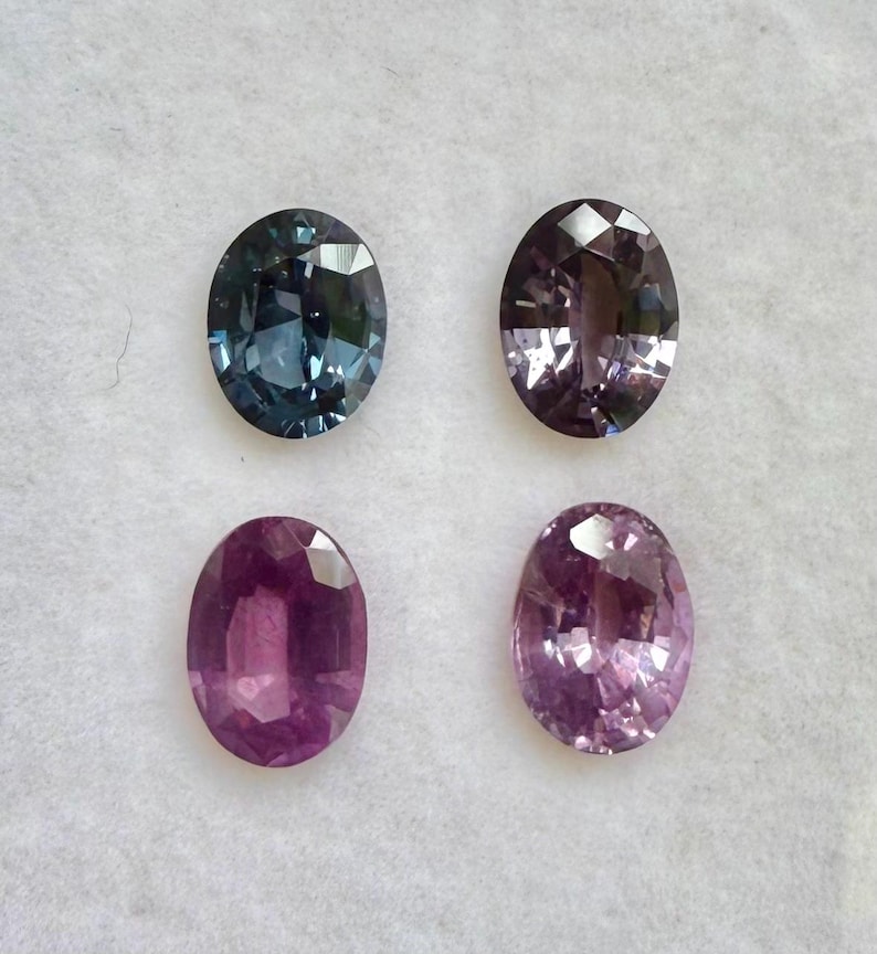 May include: Four oval-cut gemstones in shades of blue, purple, and pink are displayed against a white background. The gems vary in color intensity, with facets catching the light. These gemstones are suitable for jewelry making.