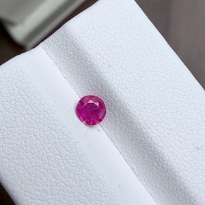 Può includere: Una gemma di rubino rotonda e sfaccettata, con una tonalità rosso-rosata intensa. La gemma è posta su una superficie bianca e strutturata, creando un contrasto che ne esalta la brillantezza. La pietra preziosa è il punto focale, mostrando la sua chiarezza e il suo colore.