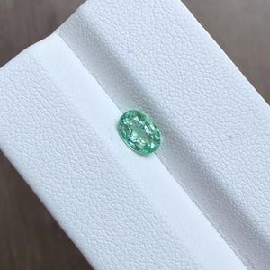 May include: A light green, oval-shaped gemstone. The gem is set against a white, textured surface. The stone has a slightly translucent appearance, with visible internal features. The overall color is a soft, inviting green.