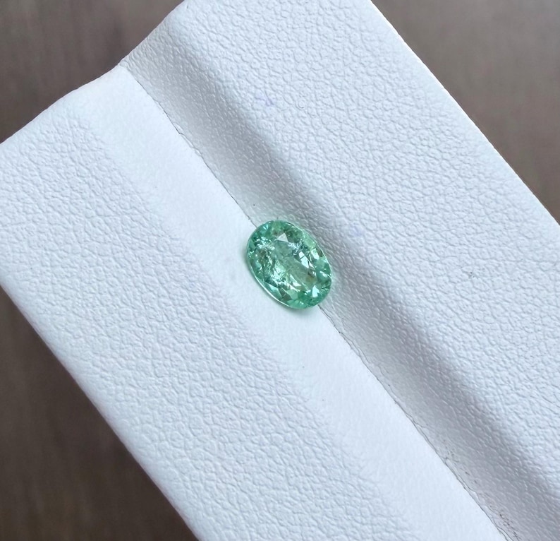 May include: A light green, oval-shaped gemstone. The gem is set against a white, textured surface. The stone has a slightly translucent appearance, with visible internal features. The overall color is a soft, inviting green.