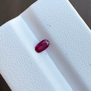 May include: A vibrant, oval-cut ruby gemstone. The deep red gem is displayed against a white, textured surface. The ruby's facets catch the light, showcasing its brilliance and color.