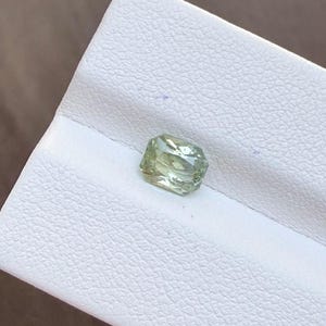 May include: A light green gemstone with a rectangular shape and faceted surfaces. The gem is set against a white background. The gemstone has a slight yellow hue and appears to be transparent.