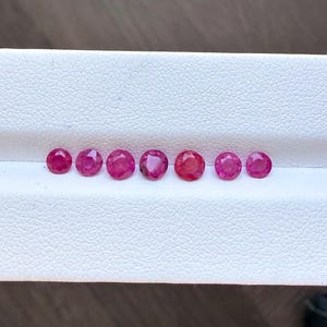 Puede incluir: Siete gemas rojas redondas y facetadas dispuestas en fila sobre un fondo blanco. Las gemas, probablemente rubíes, muestran una gama de tonos rojos, desde el carmesí intenso hasta un rojo más brillante. Cada gema está cuidadosamente tallada.