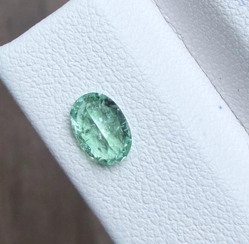 May include: An oval-shaped, light green gemstone with internal facets. The gem is set against a white, textured surface. The stone's color is a soft, translucent green, and it appears to be well-polished.