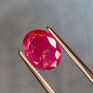 May include: A faceted, oval ruby gemstone held by tweezers. The ruby is a deep red colour with internal inclusions. The tweezers are silver-coloured and hold the gemstone against a blurred, light blue and grey background.