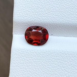 May include: A faceted, oval-shaped gemstone with a deep red hue. The gem is set against a white textured surface, showcasing its clarity and brilliance. This gemstone is ideal for jewelry making.
