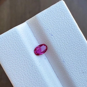May include: A vibrant, oval-cut ruby gemstone. The ruby is a deep, rich red color and is set against a white textured surface. The gemstone is displayed in a well-lit environment, highlighting its clarity and brilliance.
