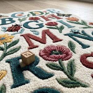 May include: A cream-colored alphabet rug with embroidered letters, flowers, and insects. The letters are in various colors, including teal, red, and brown. A wooden block with a star is on the rug. The rug has a floral border.