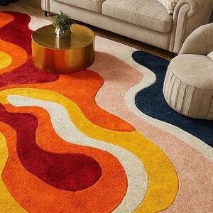 May include: A colorful area rug with an abstract design. The rug features flowing shapes in shades of red, orange, yellow, white, pink, and navy blue. The rug is placed on a wooden floor, with a sofa and chair in the background.