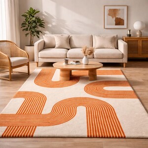 May include: A cream-colored area rug with a bold orange geometric design. The rug features curved lines and shapes, creating a modern and stylish look. The rug is placed in a living room setting with a sofa, coffee table, and other decor.