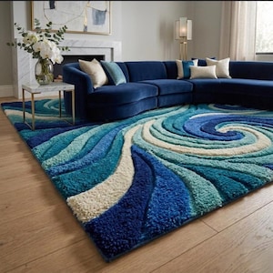 May include: A large, plush area rug with a swirling wave design in shades of blue and white. The rug is placed in a living room with a navy blue sectional sofa and a marble-topped side table. The rug's texture adds depth and visual interest to the space.