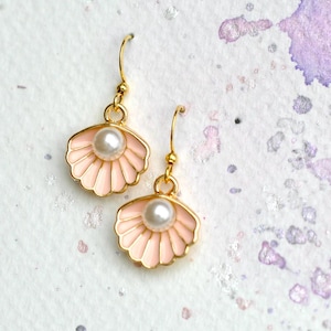 May include: Two gold-coloured seashell earrings, each with a pearl centre. The shells are pale pink with gold edging. The earrings are set against a white backdrop with purple and silver splashes.