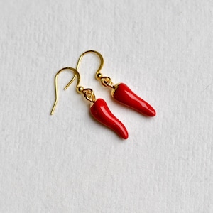 Op de afbeelding: Een paar goudkleurige oorbellen met helderrode chilipeperbedels. De oorbellen hebben een eenvoudig haakontwerp. De chilipepers zijn glanzend en gedetailleerd, tegen een witte achtergrond.