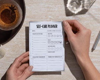 May include: A white self-care planner with black text, held by hands. The planner includes sections for daily activities, mood, water balance, and sleep. The title "SELF-CARE PLANNER" is at the top.