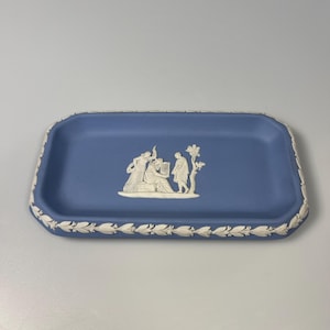 Puede incluir: Una bandeja rectangular de cerámica azul con un diseño en relieve blanco. La bandeja presenta un relieve blanco central de figuras y un borde decorativo de hojas blancas alrededor de los bordes. La bandeja es de color azul claro.