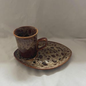May include: A brown ceramic coffee cup and saucer set. The cup has a handle and a speckled design in shades of brown and tan. The saucer is an irregular shape and has a matching speckled pattern. The cup has a slightly flared rim.