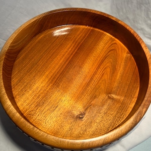 May include: A circular wooden bowl in a warm brown hue, showcasing prominent wood grain. The bowl features a smooth, polished interior and a slightly elevated rim. The exterior of the bowl has a decorative pattern.