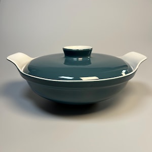 Könnte beinhalten: Eine türkisfarbene Keramik-Auflaufform mit Deckel. Die Schüssel hat zwei Griffe und einen weißen Rand. Der Deckel hat einen kleinen Knopf und einen weißen Rand. Die Schüssel steht auf einer grauen Oberfläche.