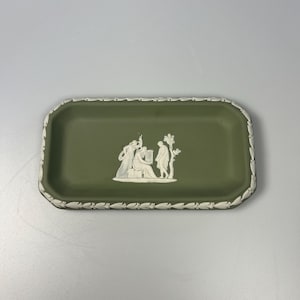May include: A rectangular, olive-green decorative tray with a white, raised relief design of classical figures. The tray has a white, leaf-patterned border. A detailed, elegant piece.