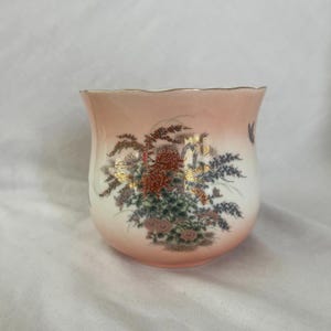 May include: A ceramic teacup with a floral design. The cup has a peach-coloured exterior that fades to white. The floral design includes red, gold, and purple flowers. The rim of the cup is trimmed with gold.
