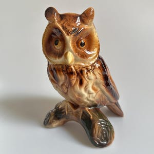 Könnte beinhalten: Eine Eulenfigur aus Keramik mit glänzender Oberfläche in Braun-, Beige- und Cremetönen. Die Eule hat große, dunkle Augen, spitze Federohren und einen hellen Schnabel und sitzt auf einem strukturierten, dunkelbraunen astförmigen Sockel.