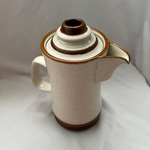 May include: A speckled ceramic coffee pot with a brown rim and lid. The pot has a handle and a spout. The lid is a tiered design with a dark brown top. The body of the pot is a light beige color with brown speckles.