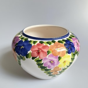 Op de afbeelding: Een ronde keramische kom met een witte basis, voorzien van een handgeschilderd bloemmotief in tinten blauw, roze, oranje, geel en paars, geaccentueerd met groene bladeren. De rand is afgewerkt met een donkerblauwe band aan de bovenzijde.
